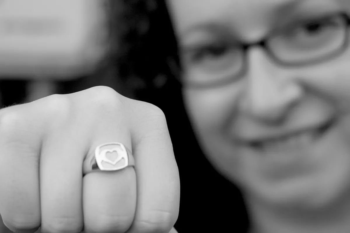 Black and white close-up of a silver ring with a heart held in two hands, worn on a raised hand, with a smiling person in glasses blurred behind